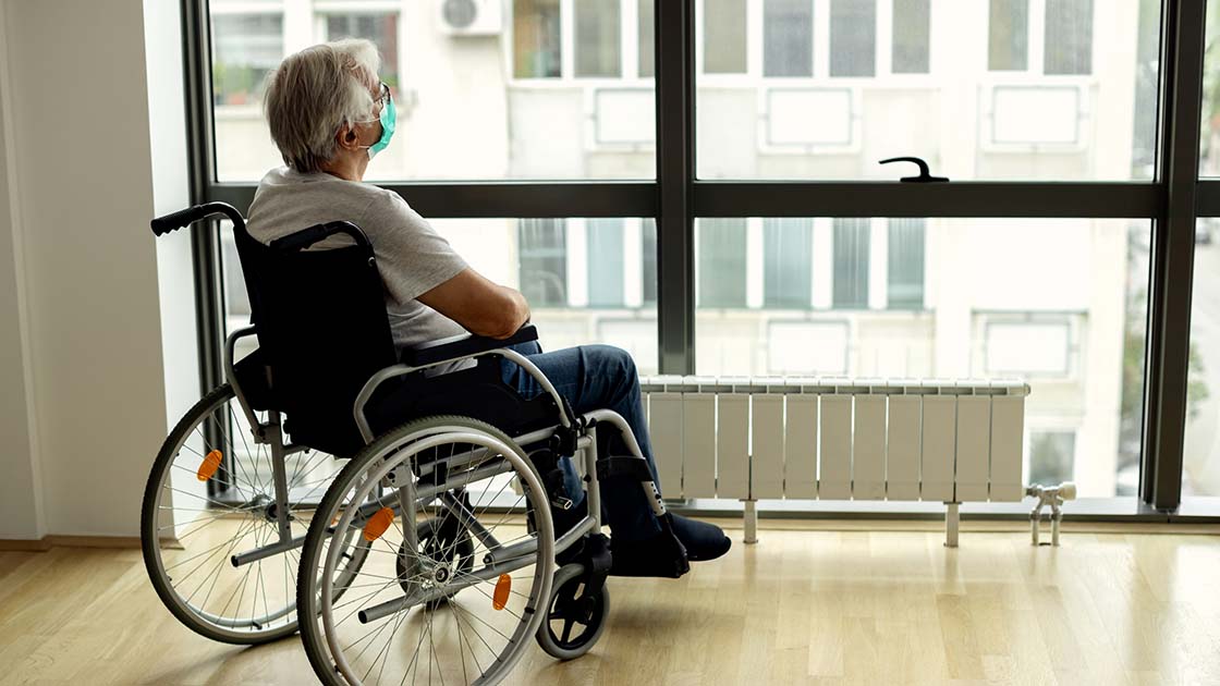 Pensive mature man in wheelchair wearing protective face mask while looking through the window during home quarantine.