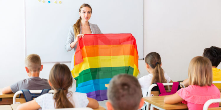 Young progressive female teacher discussing with preteen children about LGBT social movements in classroom, holding rainbow flag